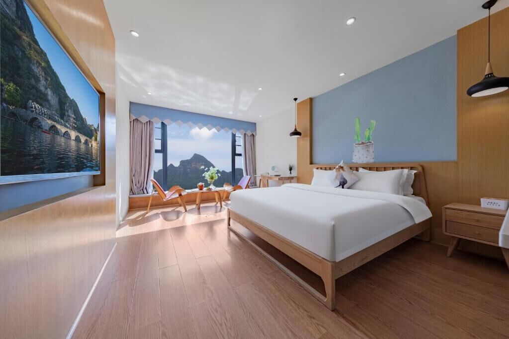 Una habitación de hotel moderna con una cama grande, piso de madera, vistas a un paisaje montañoso y una pequeña mesa con sillas.