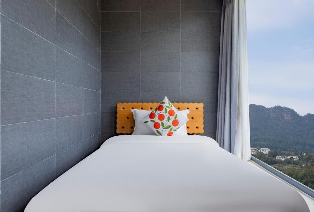 A hotel room with a bed, a wooden headboard, a pillow with a fruit pattern, and a view of mountains through a window.