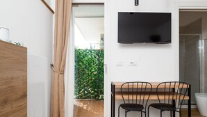 A modern kitchen with a wooden table and chairs, a flat-screen TV mounted above the table, and a sliding glass door leading to a green outdoor area. - Flowers Room – Nature-Inspired Elegance with a Private Balcony (Piano di Sorrento)