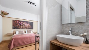 A bedroom with a bed, a painting, a wooden floor, a stone wall, and a bathroom with a large sink and mirror. - Flowers Room – Nature-Inspired Elegance with a Private Balcony (Piano di Sorrento)