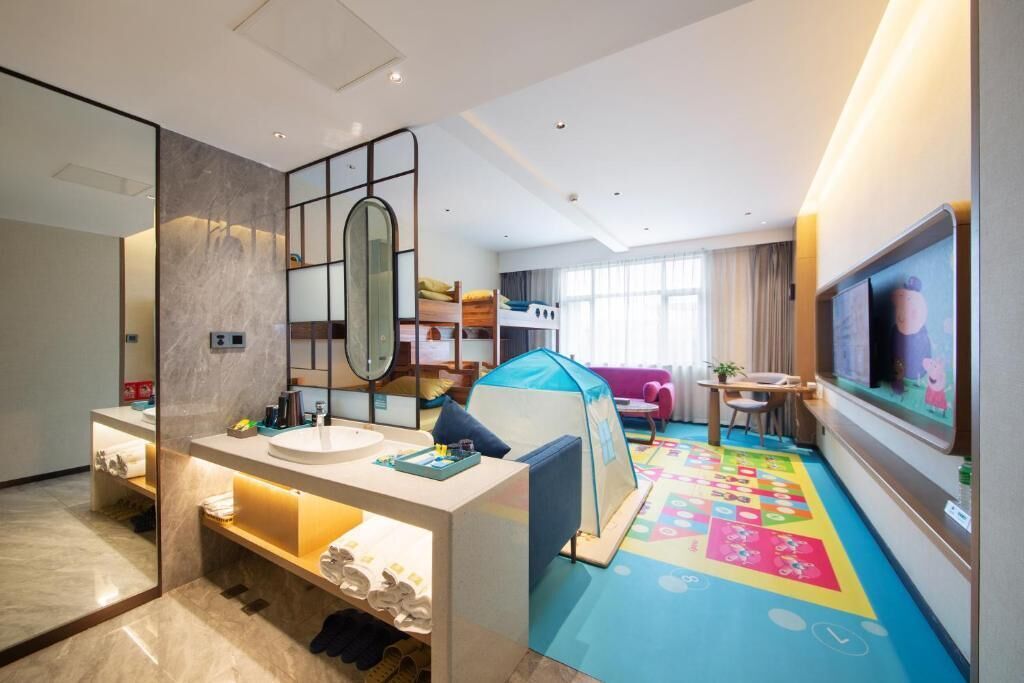 Habitación de hotel con un espejo grande, una cama individual, una zona de juegos con colchoneta, televisor y una pequeña mesa con silla.