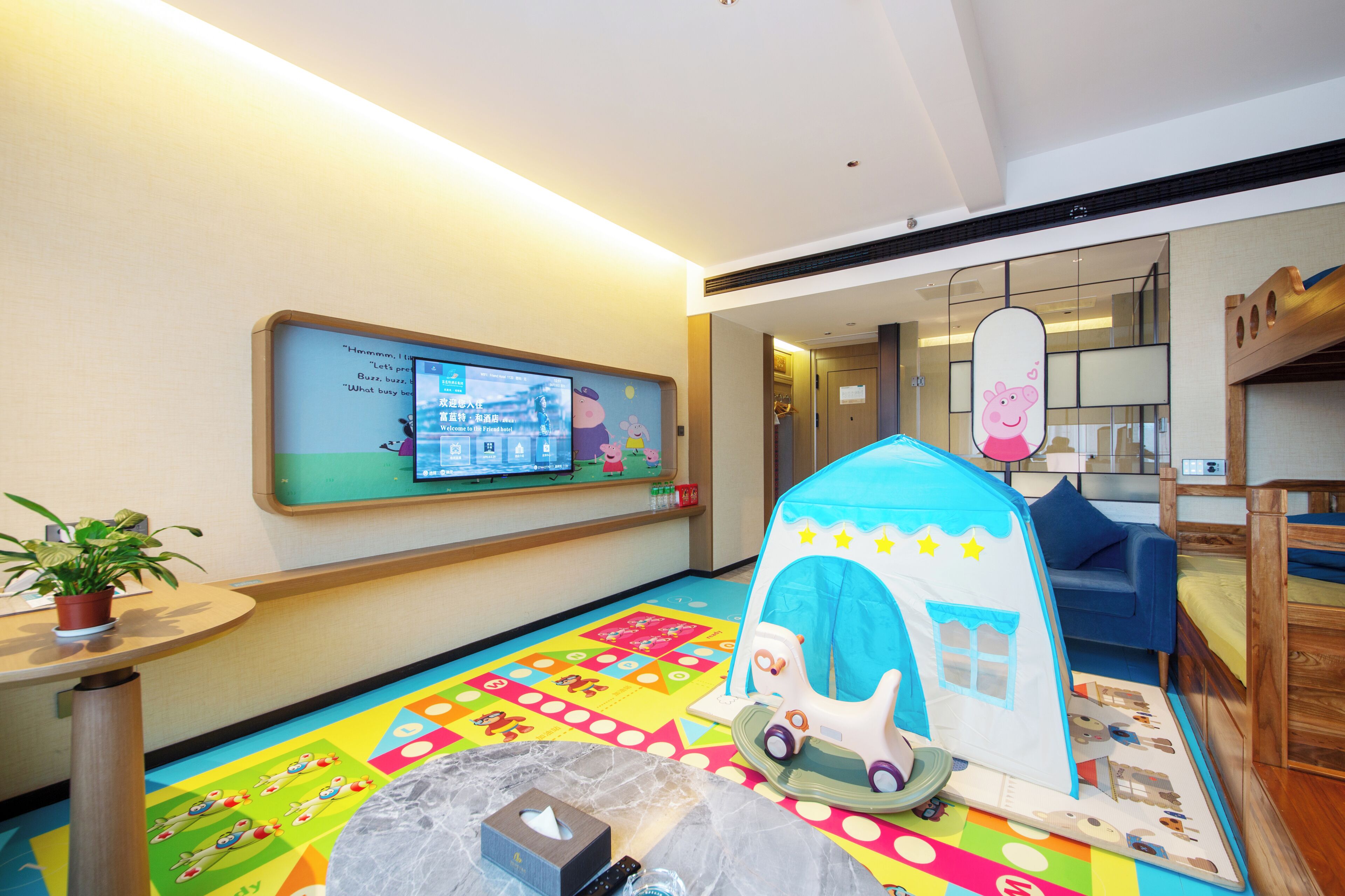 Un espace de jeux pour enfants avec un sol coloré, une structure de jeu en forme de tente et un écran de télévision diffusant des dessins animés.