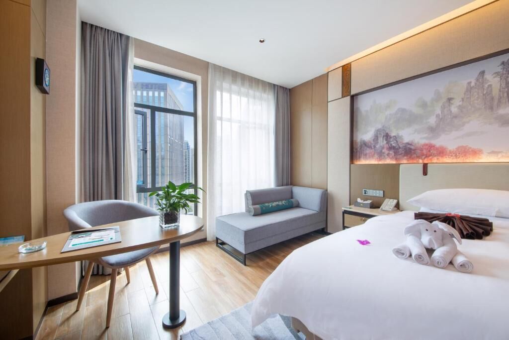 A hotel room with a bed, a desk, a chair, a sofa, a painting, and a window with curtains.