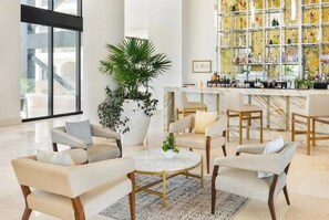 Un espace de détente moderne, avec des canapés, une table basse, une grande plante en pot et un bar doté d’étagères remplies de bouteilles.