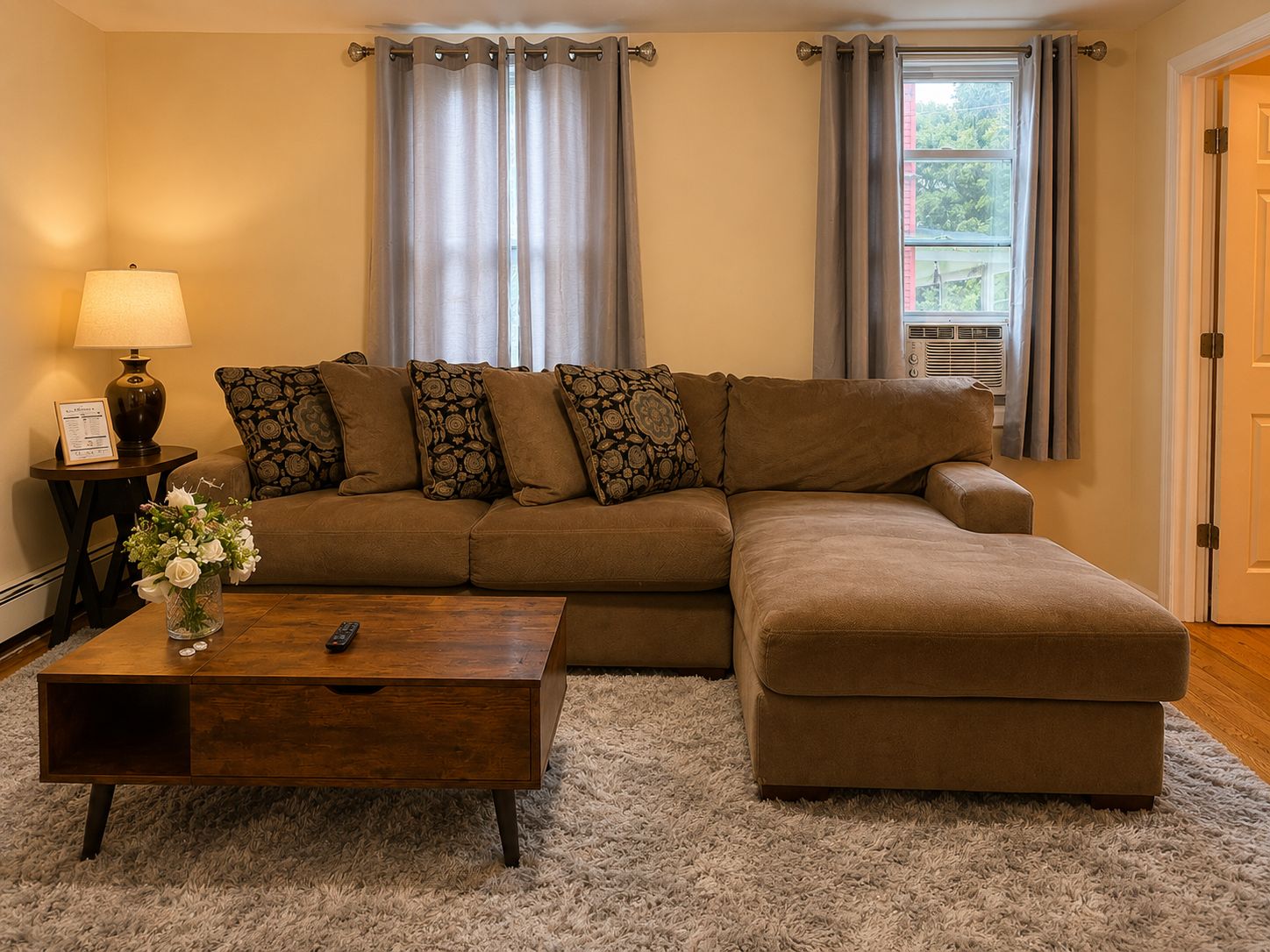 A living room with a brown sectional sofa, a wooden coffee table, a lamp, and a vase with flowers.
