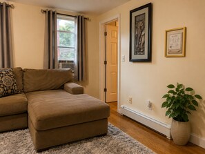 A living room with a brown sectional sofa, a patterned throw pillow, a potted plant, and a framed picture on the wall.