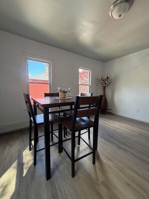 A dining area with a wooden table and chairs, two windows with views, and a vase with dried flowers.