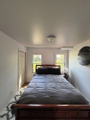 A bedroom with a bed, two windows, a door, and a wall-mounted lamp.