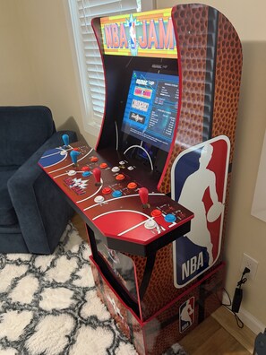 A basketball arcade game machine with a screen displaying NBA JAM, a couch, and a patterned rug.