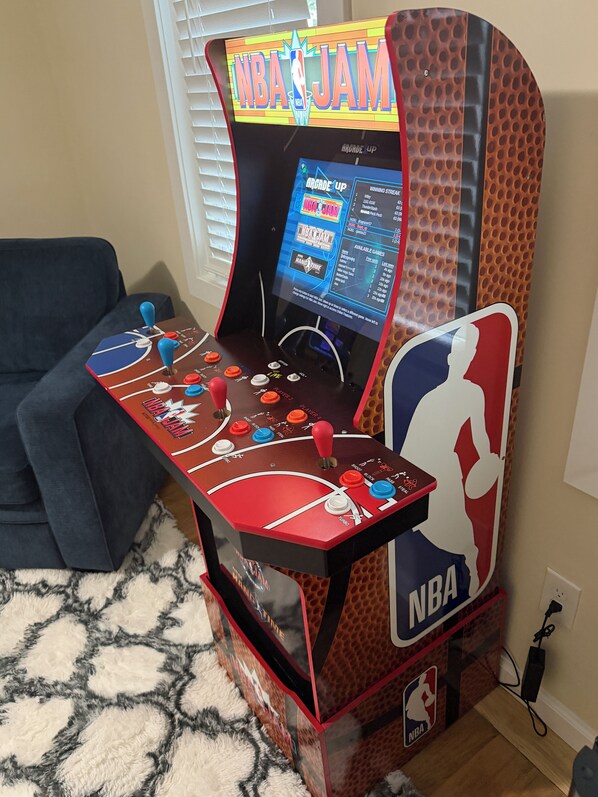 Une borne d’arcade de basket-ball avec un écran affichant le jeu NBA JAM, un canapé et un tapis à motifs.