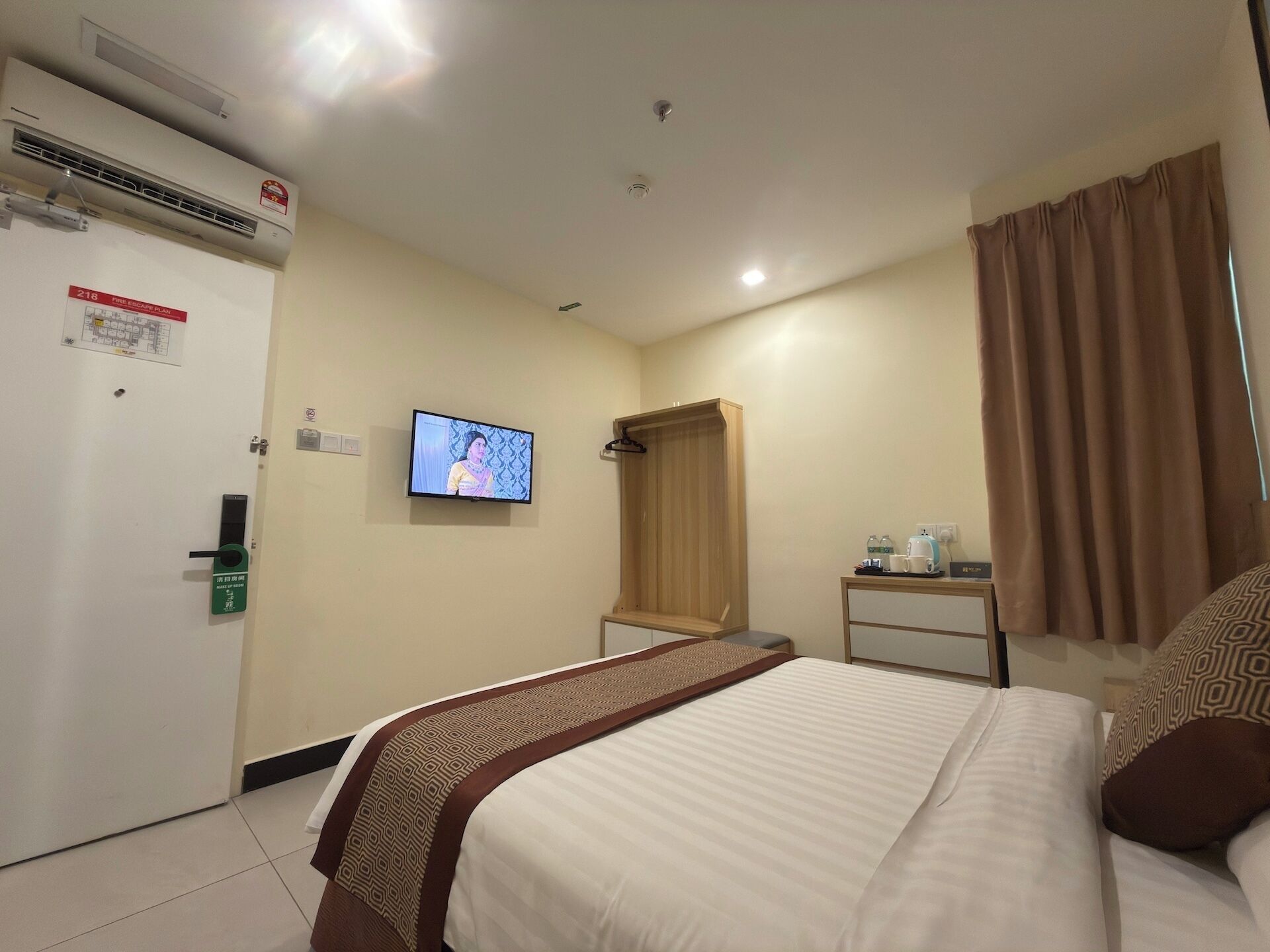 A hotel room with a bed, a television, a wardrobe, a small table with a kettle, and a door with a fire safety sign.