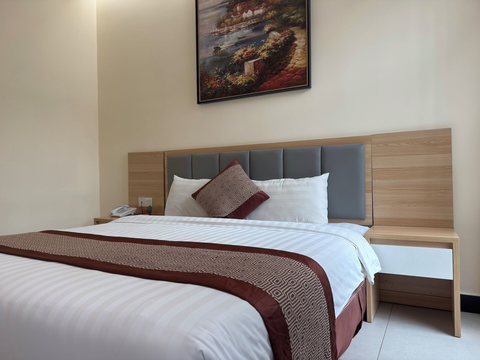 Una habitación de hotel con una cama, una mesita de noche con un teléfono, una lámpara de noche y un cuadro en la pared.