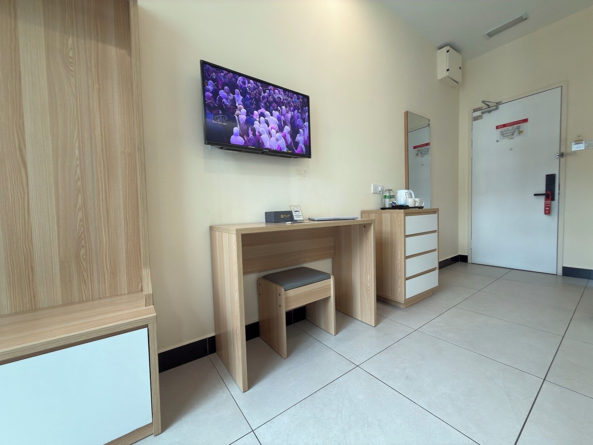 Une chambre d’hôtel avec un bureau en bois, un téléviseur fixé au mur, un miroir et une porte dotée d’une poignée rouge.