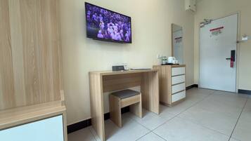 A hotel room with a wooden desk, a television mounted on the wall, a mirror, and a door with a red handle.