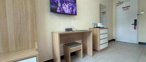 A hotel room with a wooden desk, a television mounted on the wall, a mirror, and a door with a red handle.