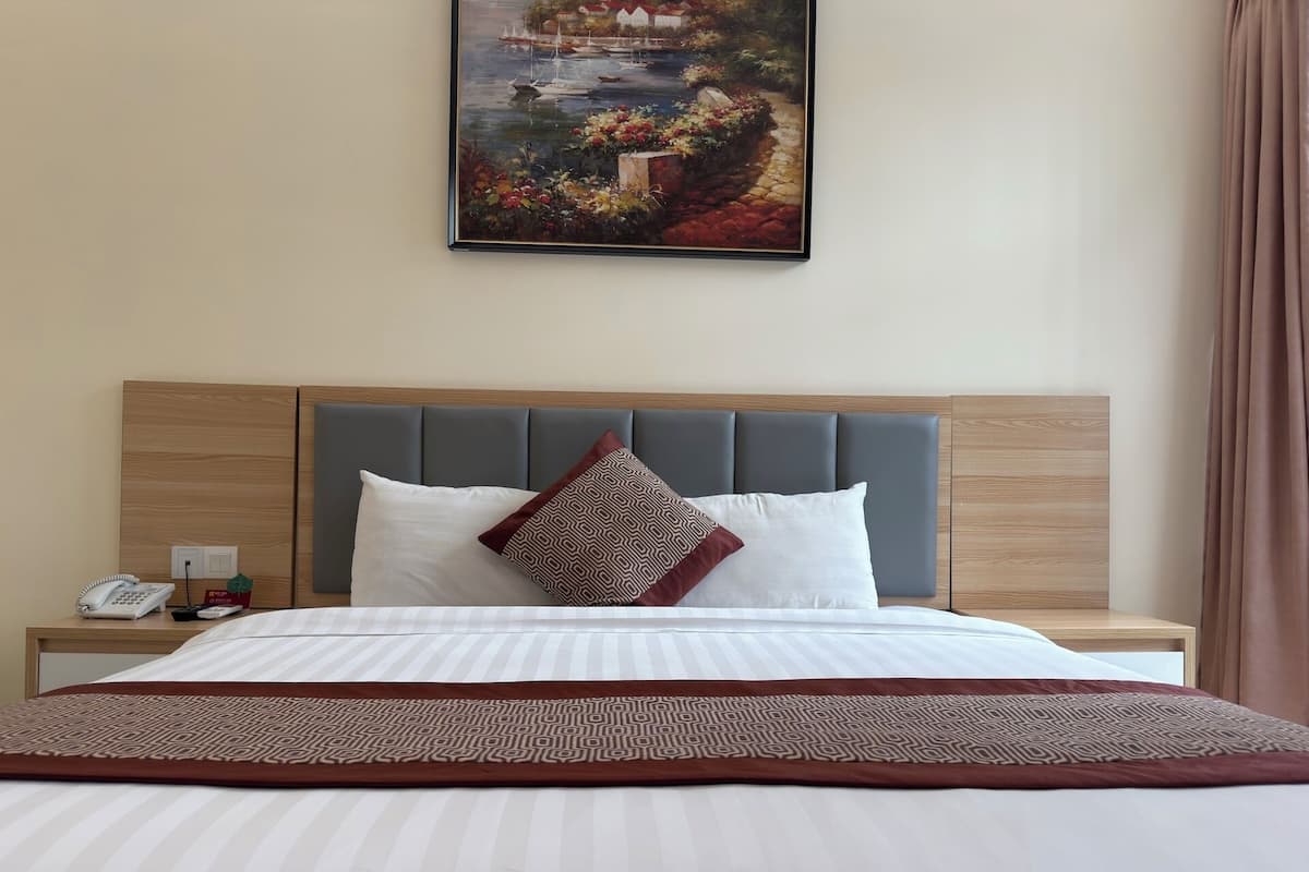Una habitación de hotel con cama, mesita de noche, teléfono, un cuadro y una cortina.