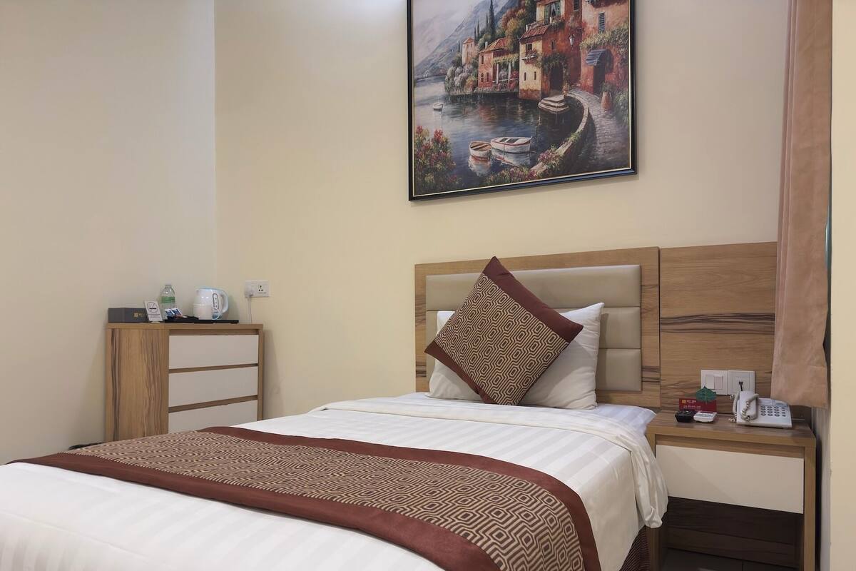 Un cuarto de hotel con una cama, una mesita de noche, un cabecero de madera, un cuadro de un paisaje lacustre y un teléfono sobre la mesita.