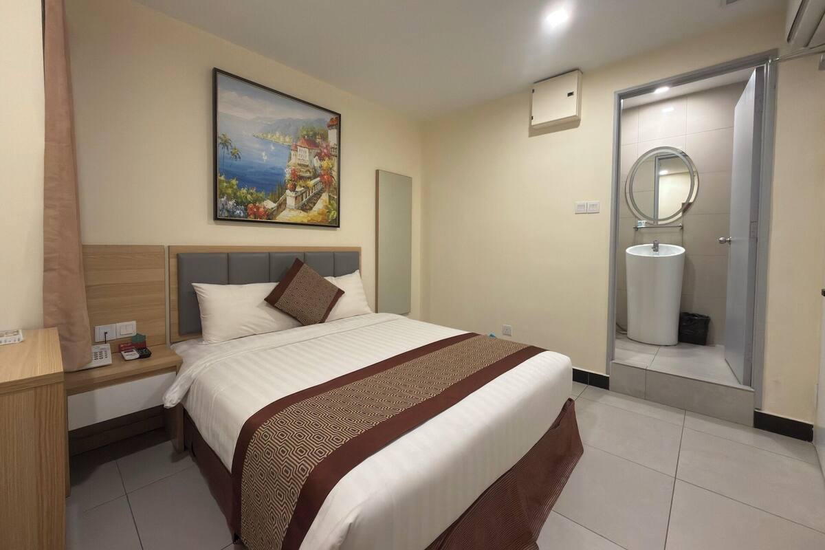 Habitación de hotel con cama, mesita de noche, un cuadro en la pared, baño con lavamanos y espejo, y una puerta.