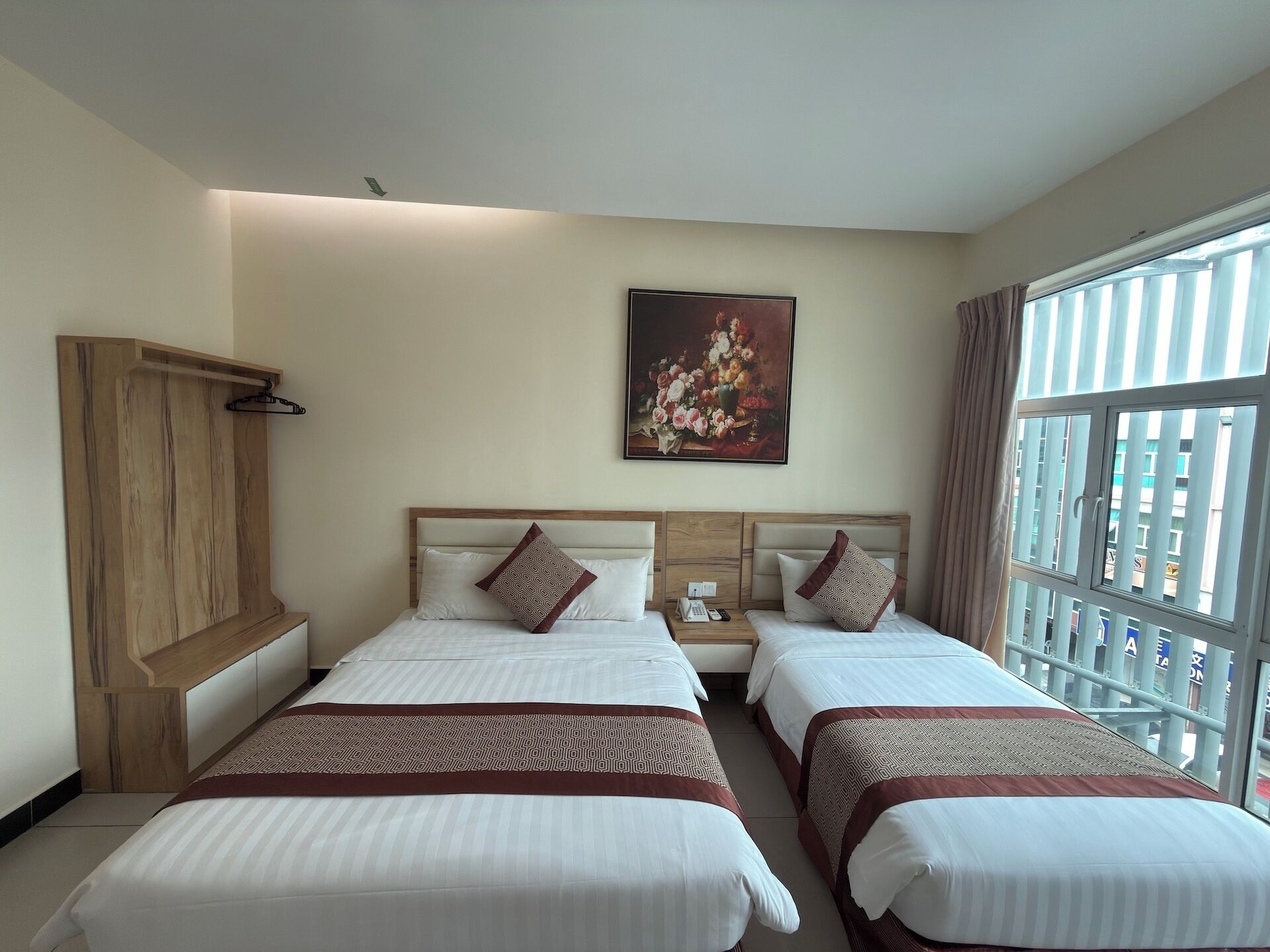 A hotel room with two beds, a wooden wardrobe, a painting on the wall, and a window with a view.