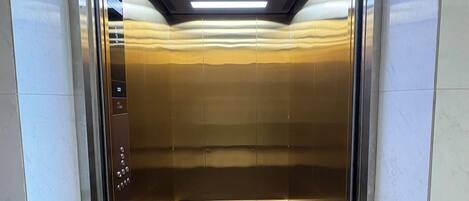 An elevator with metallic walls, a reflective floor, and a ceiling with fluorescent lighting.