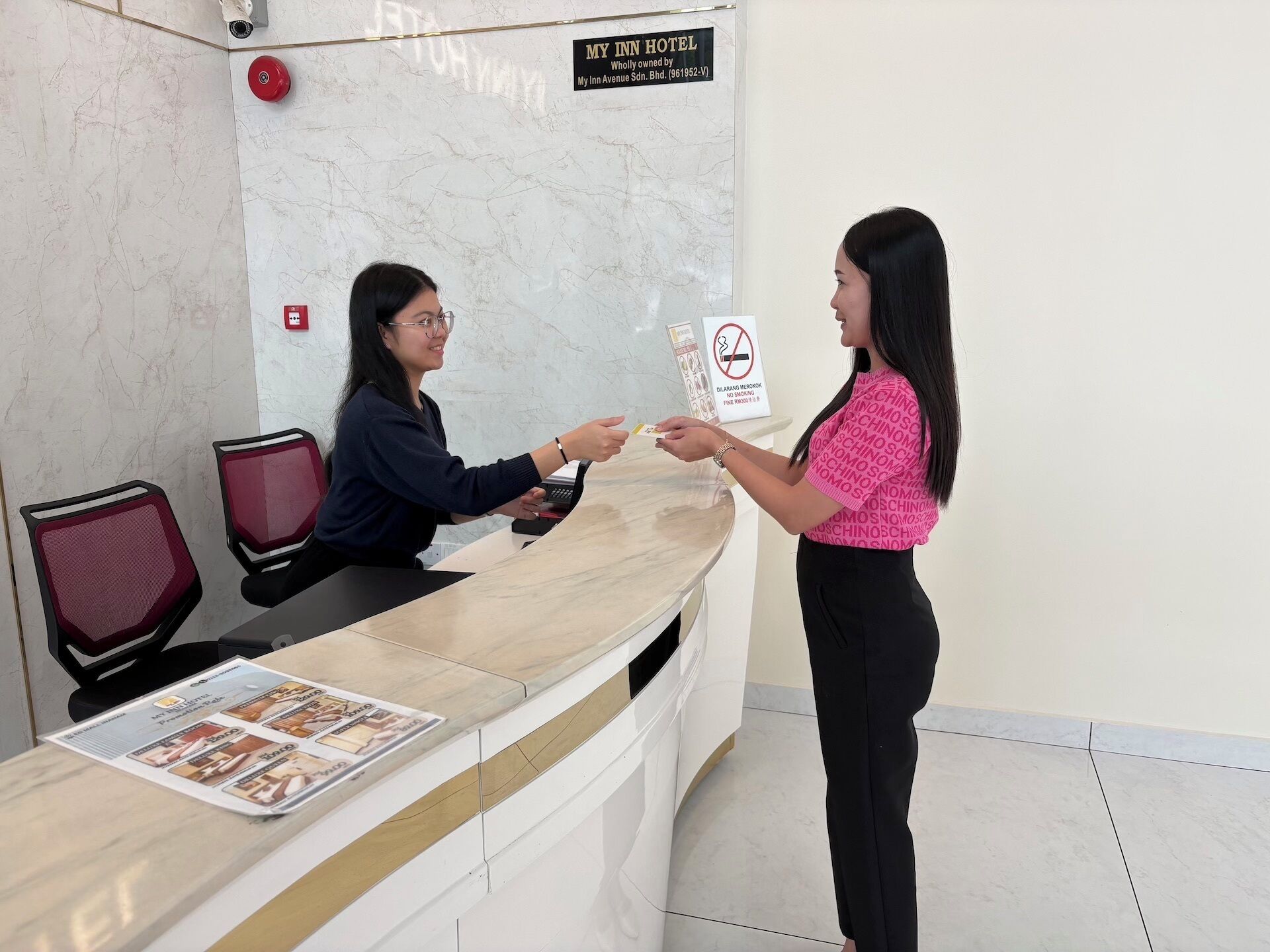 Una recepción de hotel con pared de mármol, un letrero de prohibido fumar y una recepcionista.