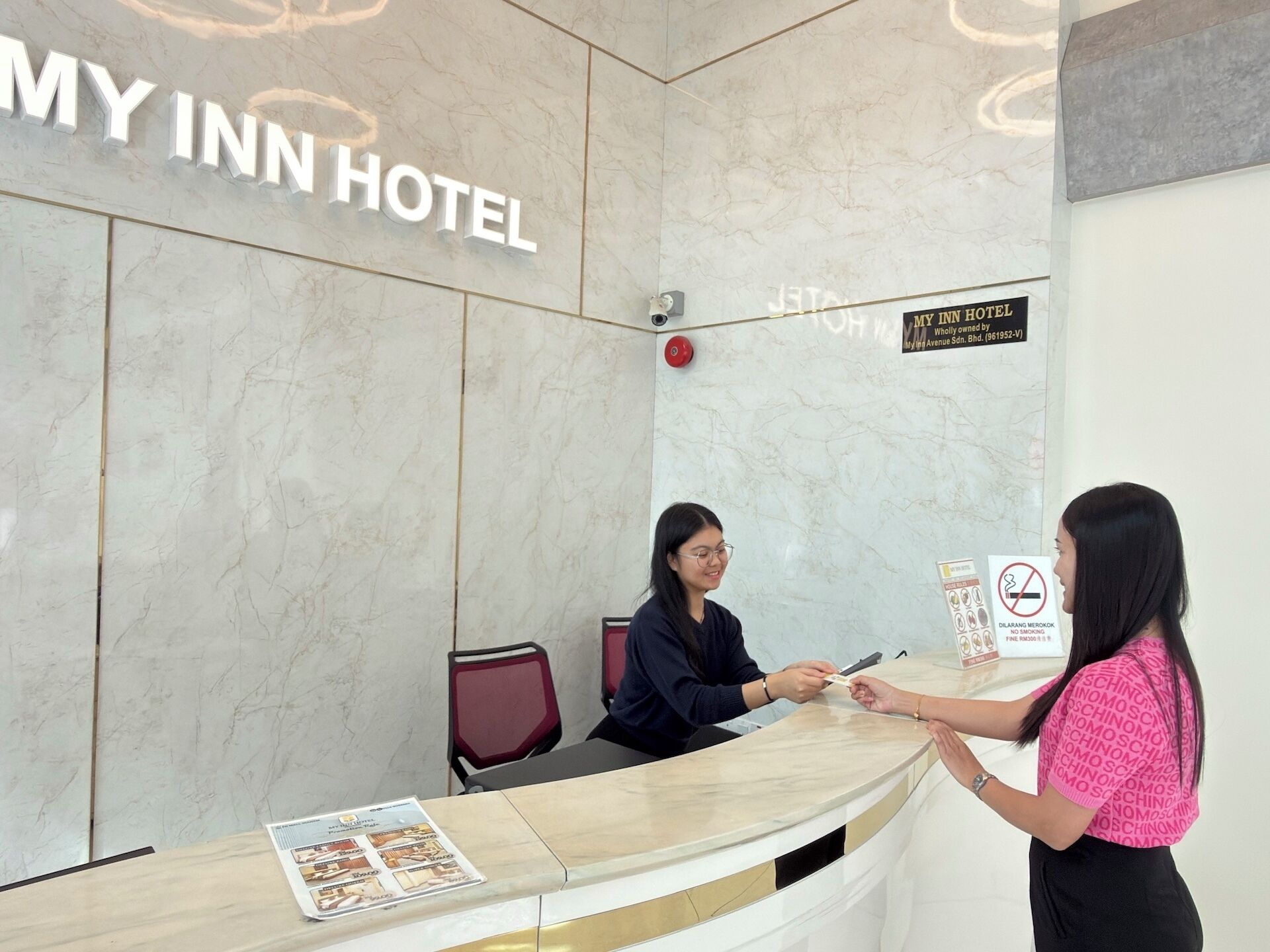 A hotel reception desk with a marble wall, a sign indicating the hotel name, and a no-smoking sign.