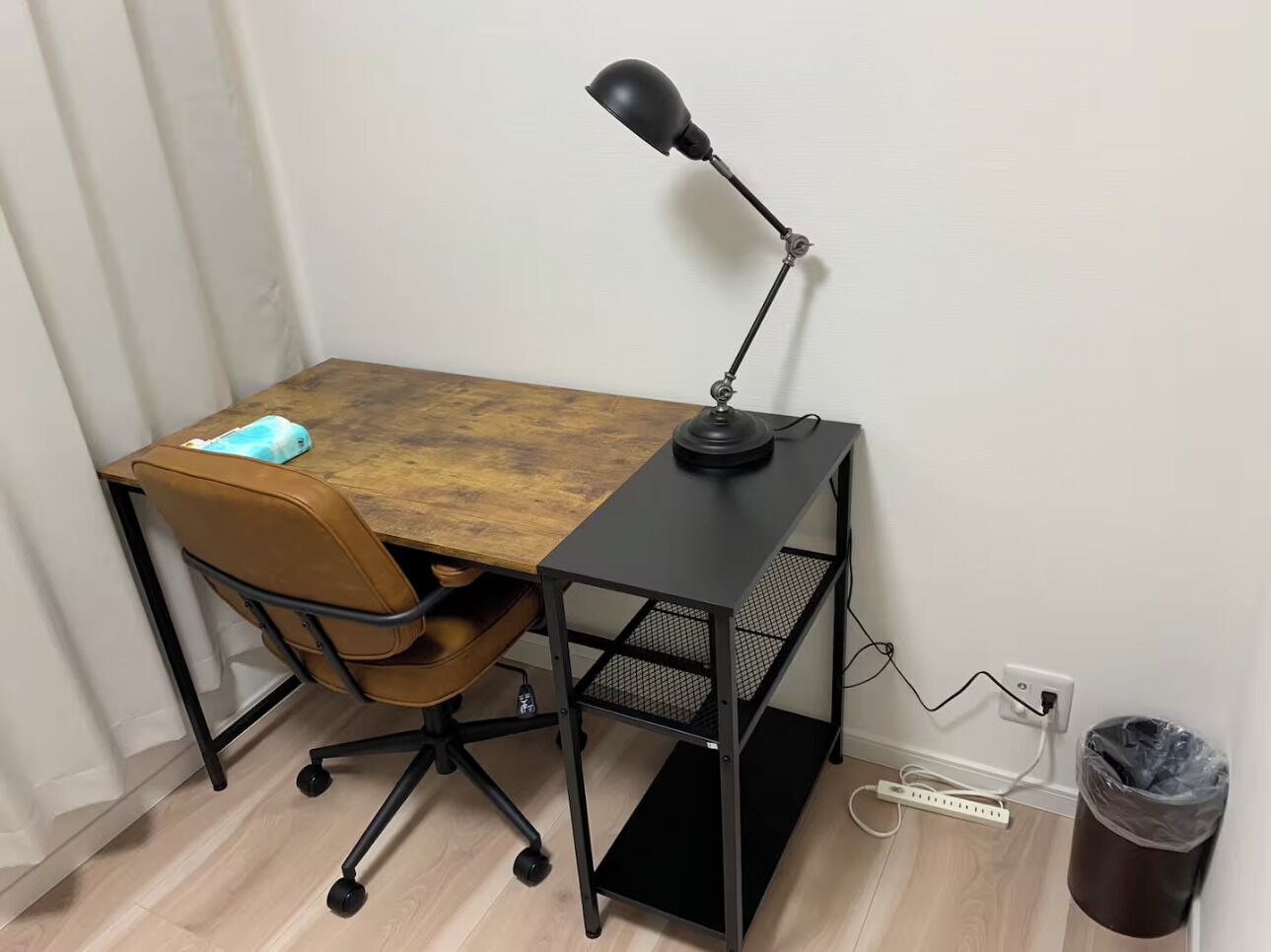 A desk with a chair, a lamp, a trash bin, and a power strip.