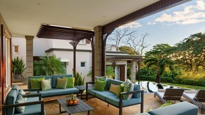 Ett takförsett utomhusområde med sittplatser, soffor och bord, en pergola och utsikt över en byggnad och grönområden.
