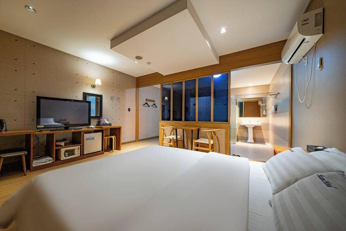 A hotel room with a large bed, a TV, a desk with a computer, a window with a view, and a bathroom with a sink and mirror.