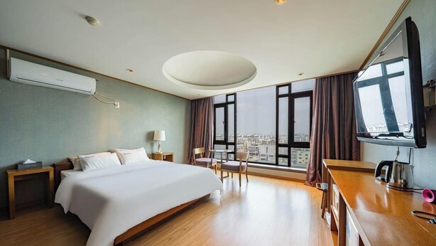A hotel room with a large bed, a desk with a chair, a TV, and a city view. - Hasen Nabeeya Hotel (Siheung)