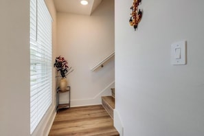 A hallway with a wooden floor, a white wall, a small shelf with a vase of flowers, and a staircase with wooden steps. - Cozy Gulfport Beach Home - Seahorse Harbor (Gulfport)