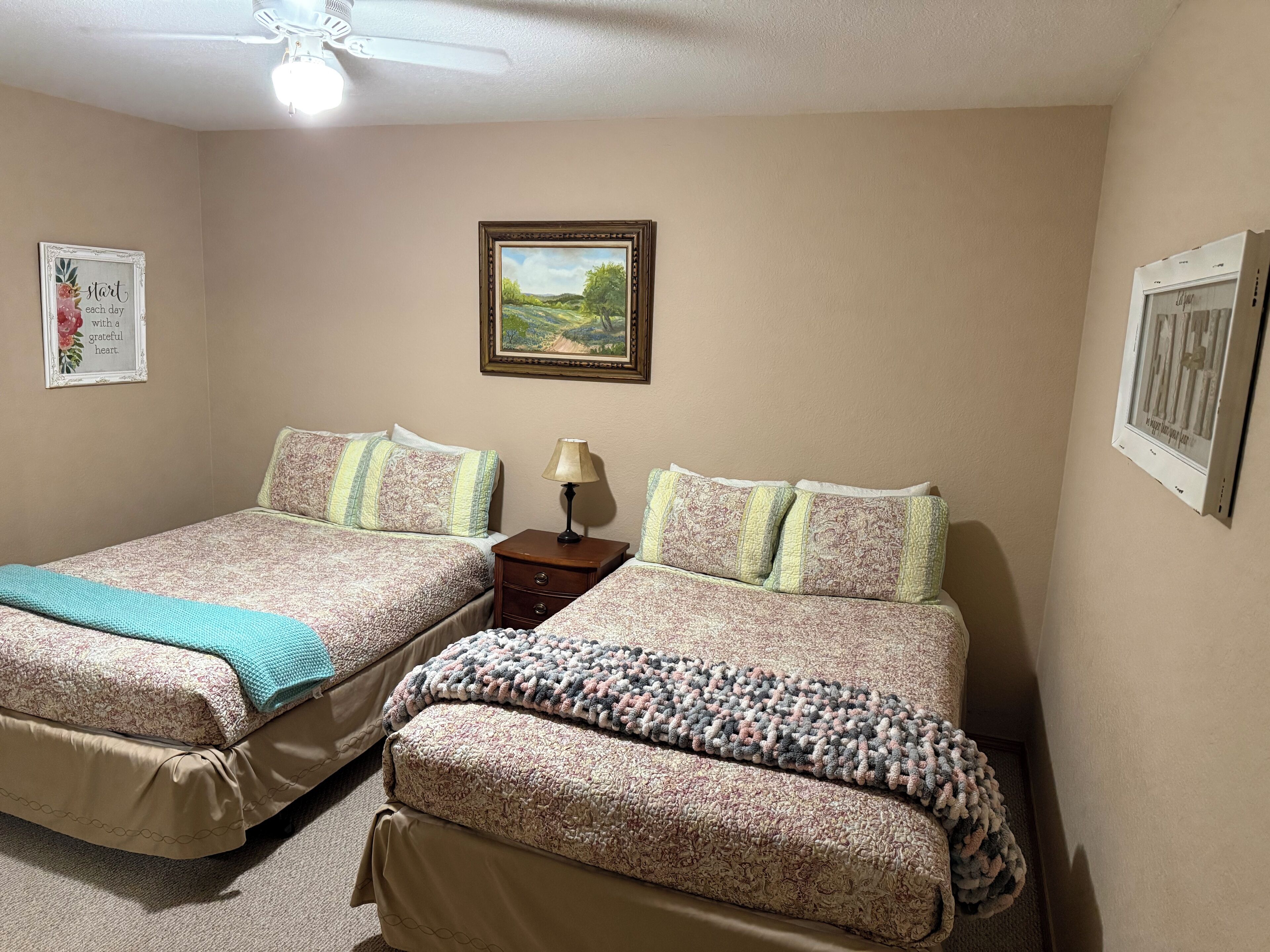A bedroom with two beds, a nightstand, a ceiling fan, and framed pictures on the walls.