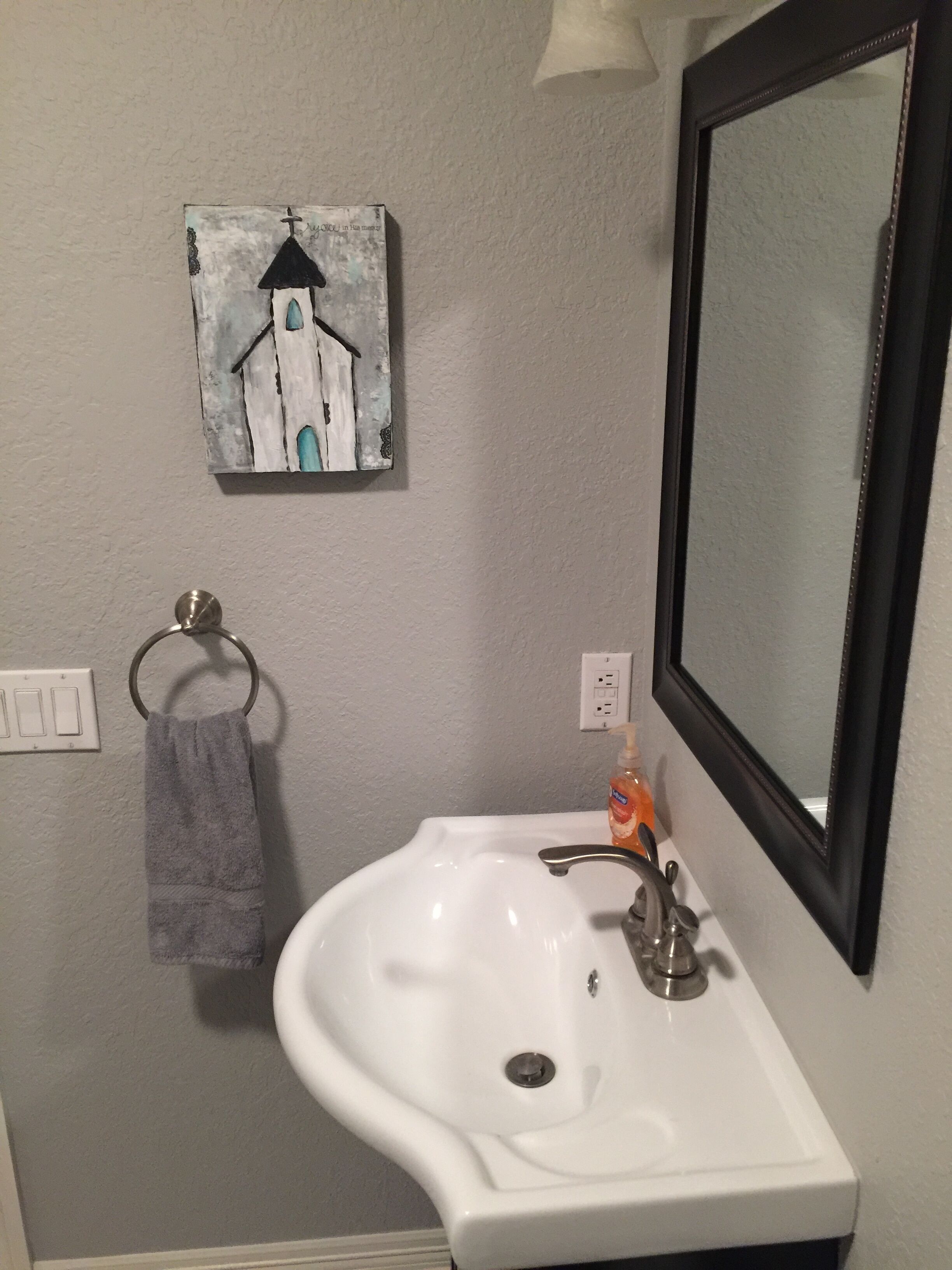 Une salle de bain avec un lavabo blanc, un miroir, un porte-serviettes et un tableau représentant une maison accroché au mur.