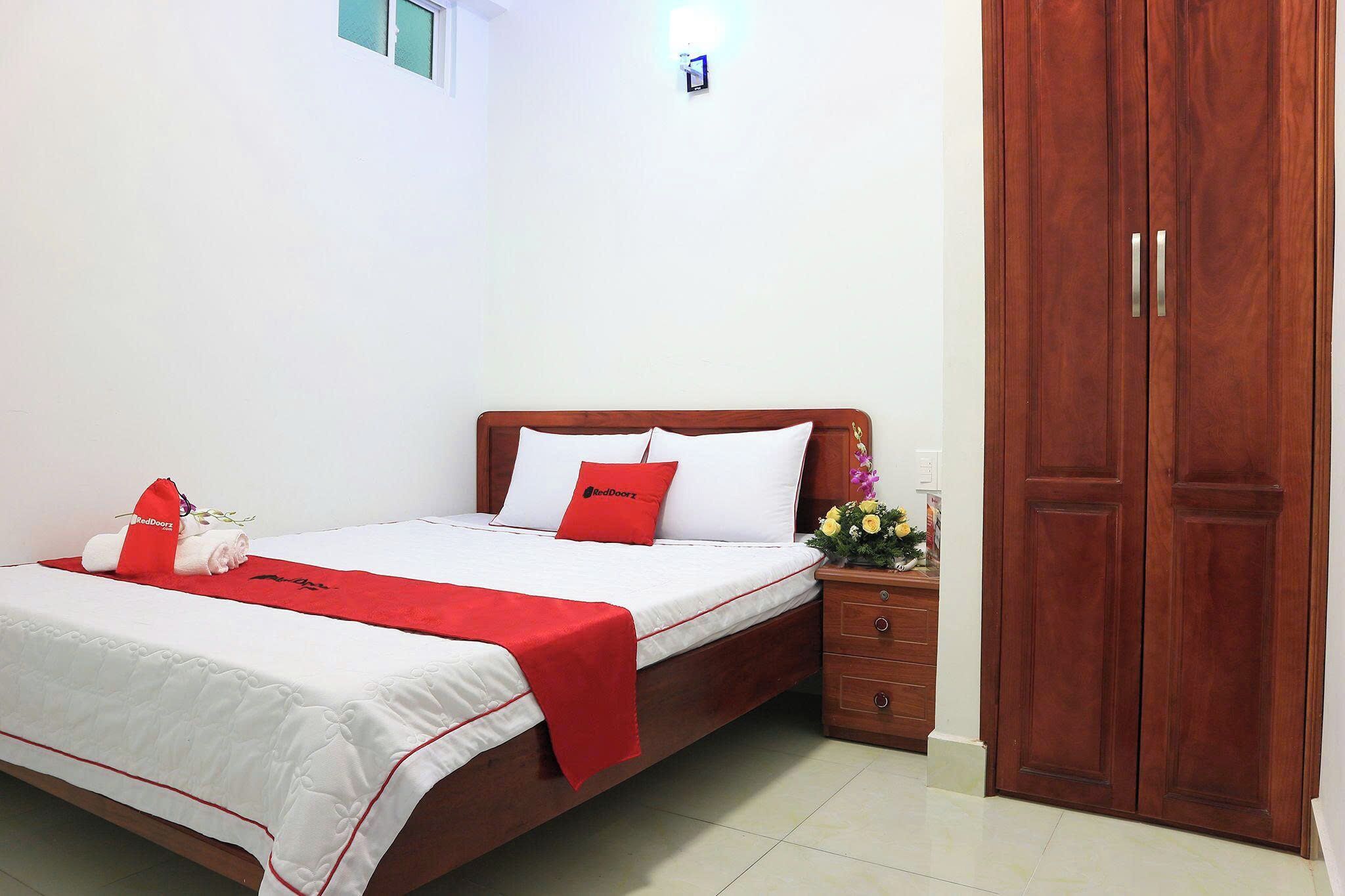 A neatly made bed with a red blanket, a wooden headboard, and a nightstand with a vase of flowers.