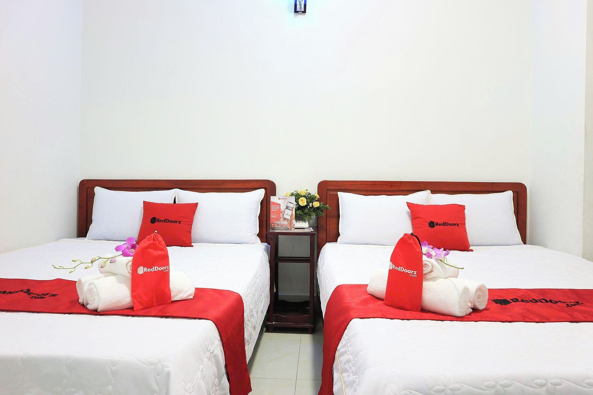 Une chambre d’hôtel avec deux lits, chacun recouvert d’une couverture et doté d’un oreiller rouge, une table de chevet avec un vase de fleurs, et le logo RedDoorz imprimé sur les oreillers et la couverture.