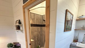 A room with a wooden floor, a cowhide rug, a small table with a plant, a framed picture, and a doorway leading to a shower area with a wooden wall. - Relaxing Rustic Barndominium with Mountain View’s in Challis Idaho (Challis)