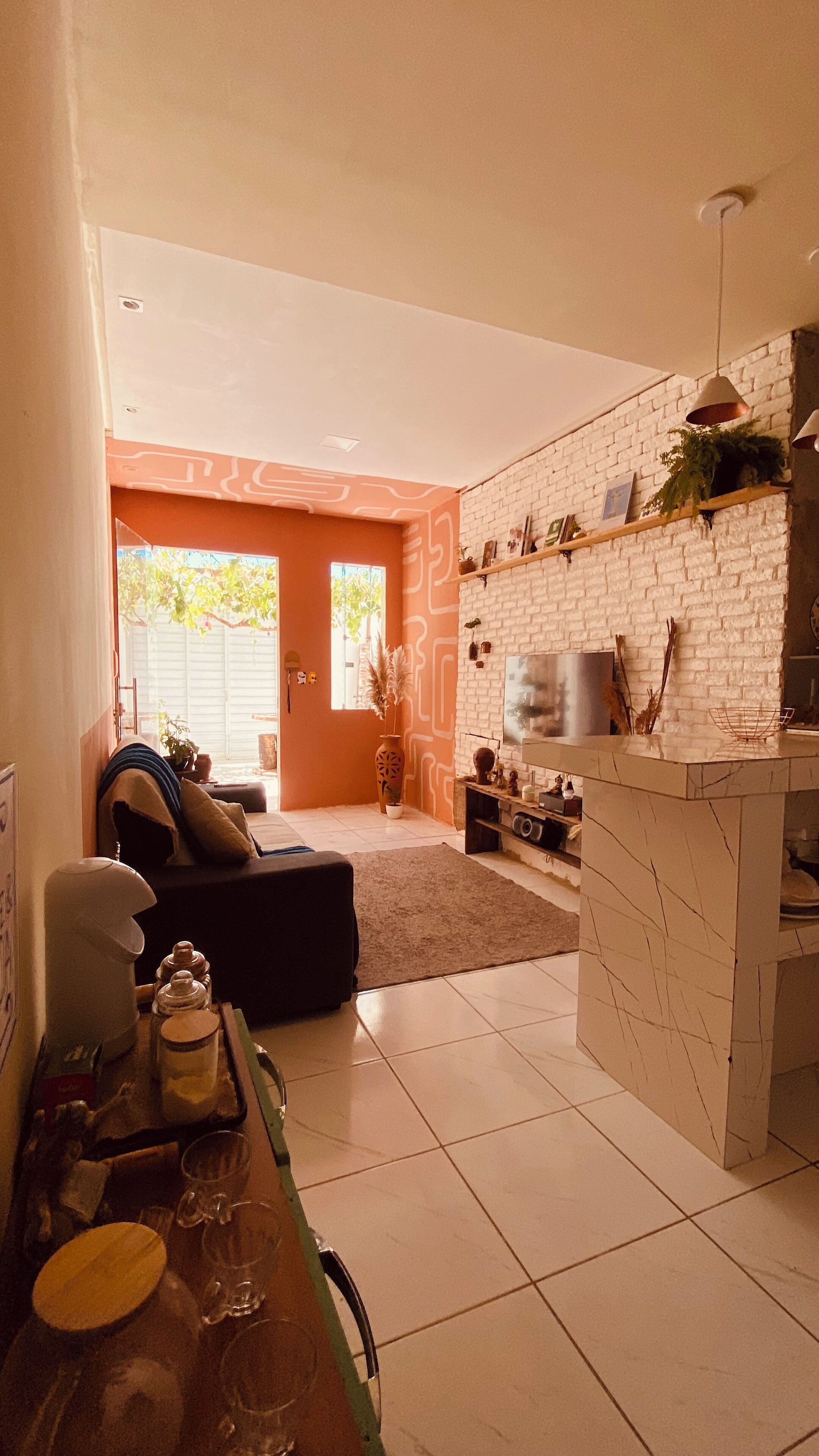 Eine moderne Küche mit weißer Ziegelrückwand, ein Essbereich mit Tisch für zwei Personen und ein Wohnzimmer mit Sofa und großem Fenster.