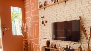 A living room with a white brick wall, a flat-screen TV mounted above a wooden console, a sofa, and a patterned rug. - Bossa Nova Home - a private house 5 minutes from the beach with Brazilian style! (Barra dos Coqueiros)