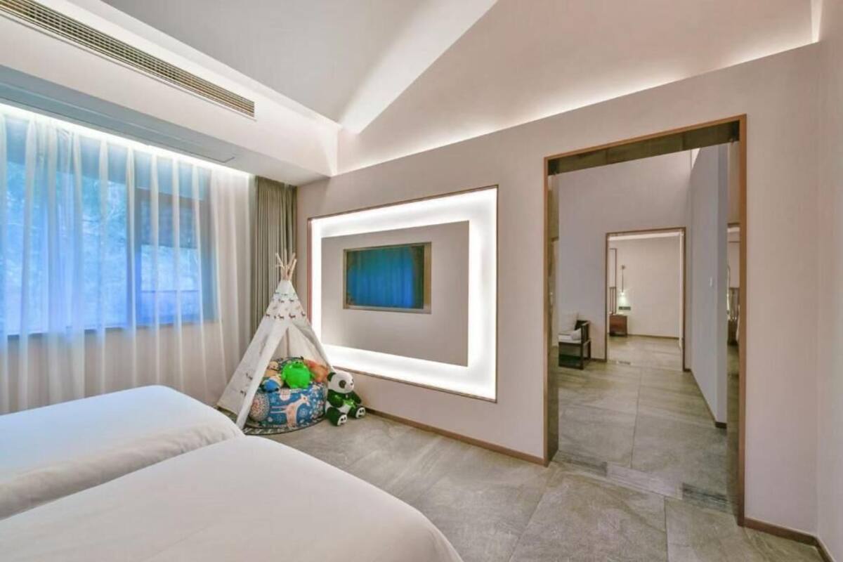 Hotellihuone, jossa on kaksi sänkyä, suuri ikkuna läpikuultavilla verhoilla, seinälle ripustettu taulu, ovi, joka johtaa toiseen huoneeseen, ja pieni leikkialue leluineen.