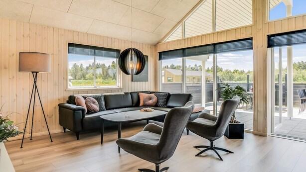 A modern interior with a sectional sofa, two chairs, a floor lamp, and large windows. - 5 Star Holiday Home in Norre Nebel (Norre Nebel)