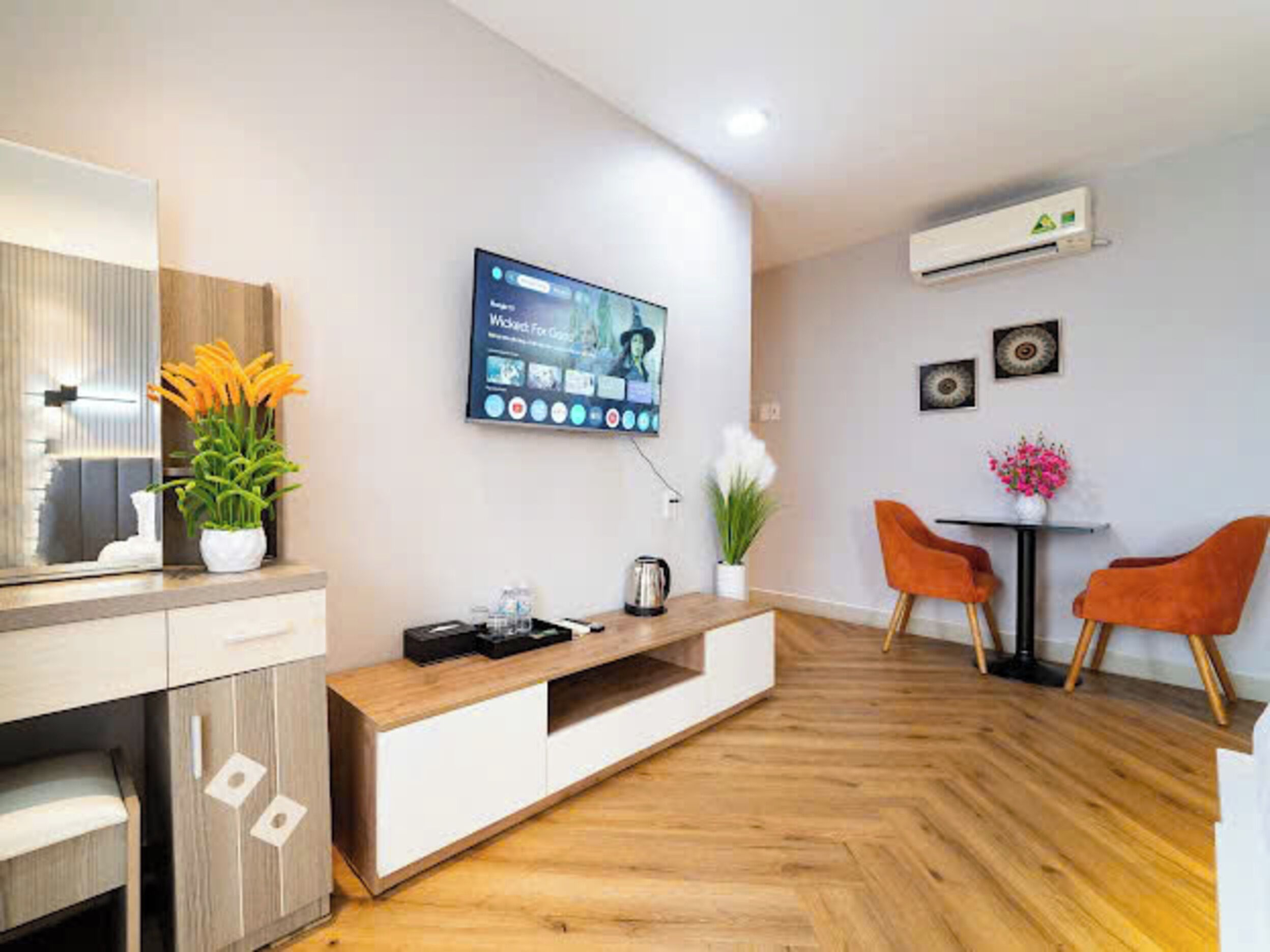 Un soggiorno moderno con una TV a schermo piatto montata al muro, un tavolino di legno, due poltrone arancioni e un piccolo tavolo con un vaso di fiori.