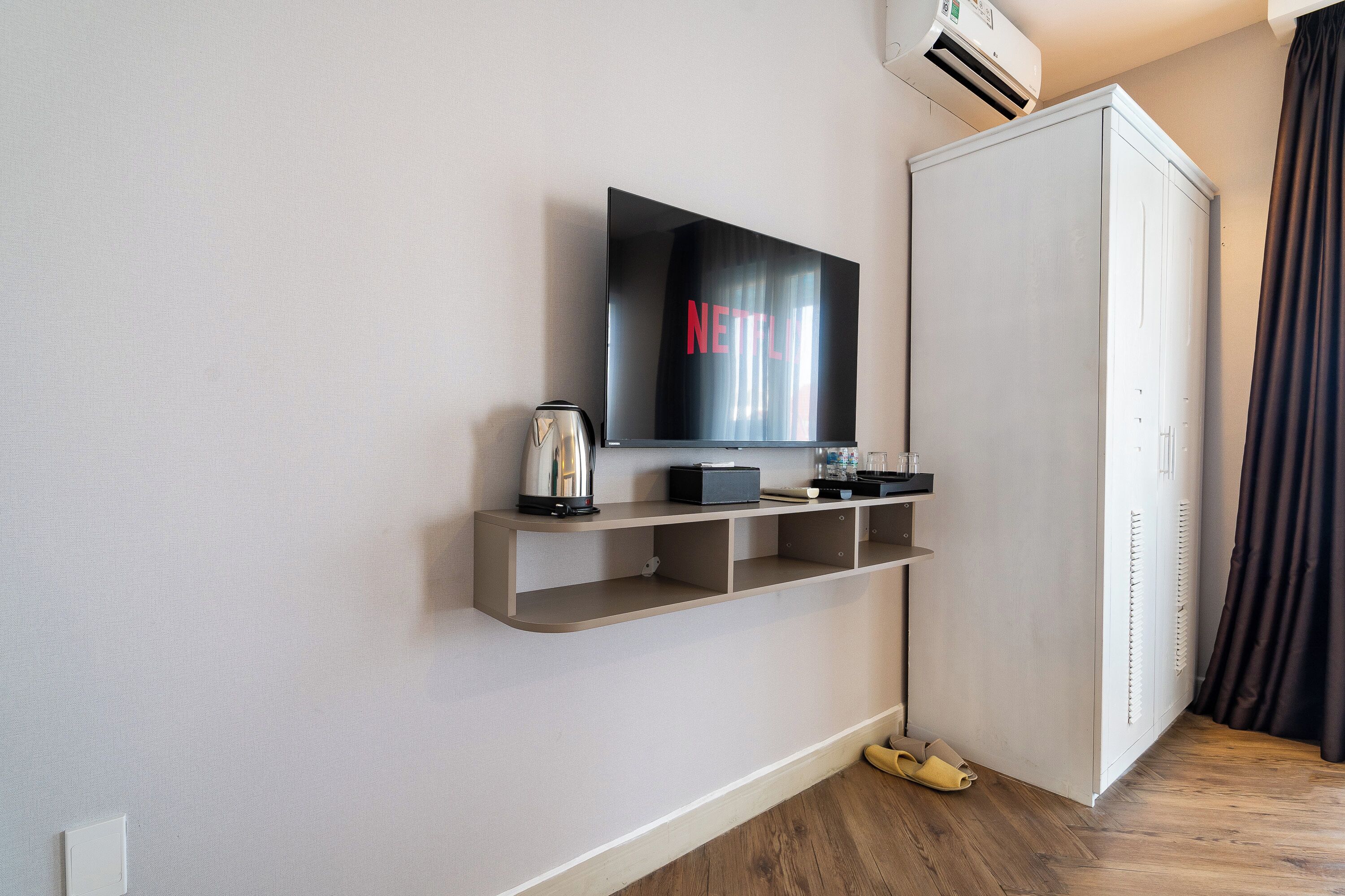 Una habitación de hotel moderna con una televisión montada, una cafetera, un estante con un control remoto, un armario y suelo de madera.