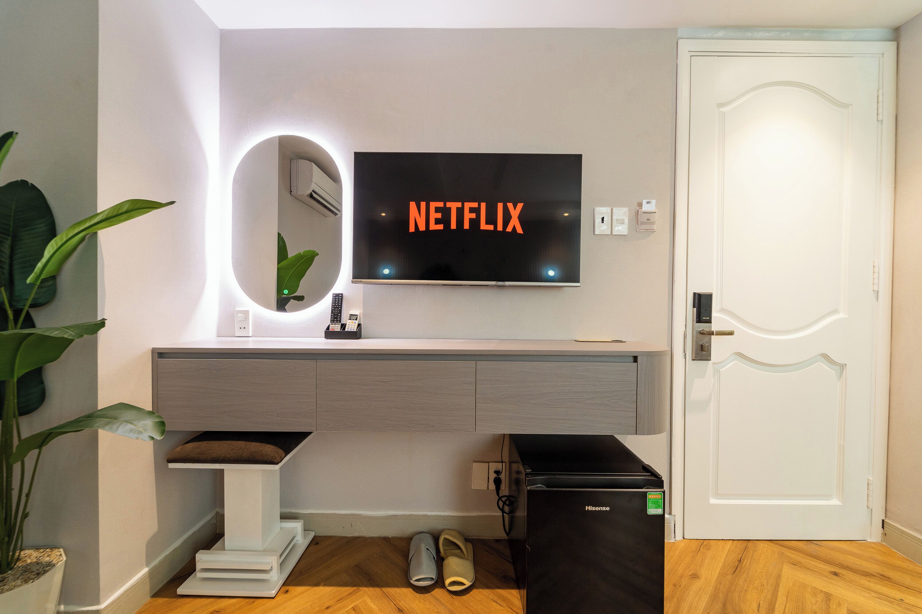 Una habitación moderna con una televisión mostrando 'NETFLIX', una puerta blanca, un refrigerador negro pequeño, una mesita blanca con un espejo y una planta en maceta.