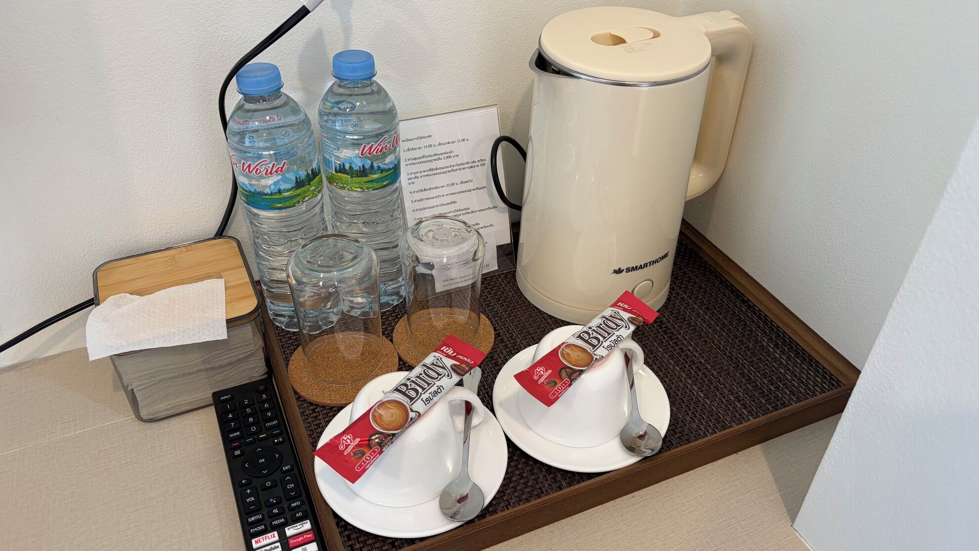 A hotel room's minibar with water bottles, a coffee maker, a remote control, a glass jar, and a packet of coffee.