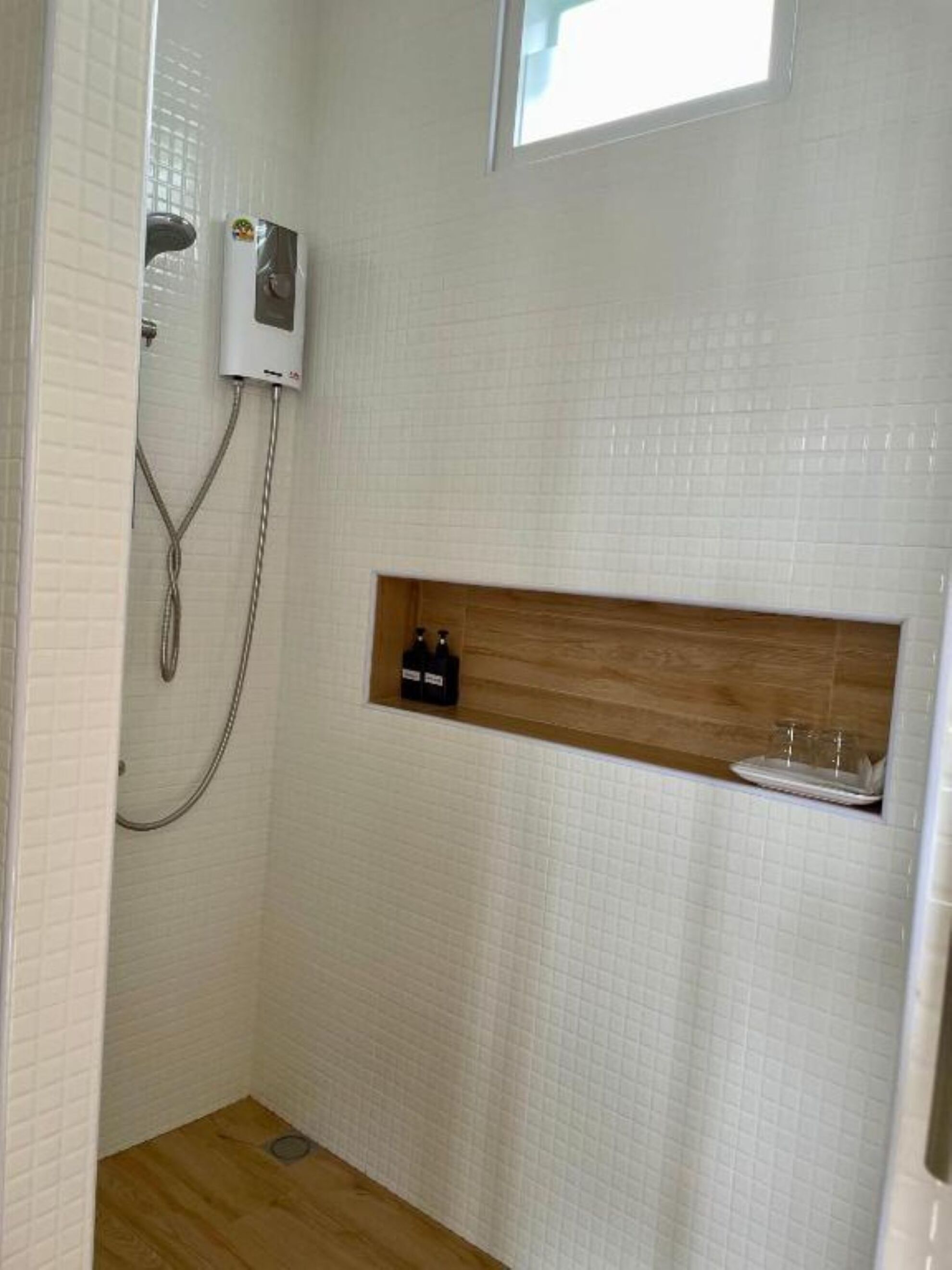 A shower with a wooden shelf, a window, and a white tiled wall.