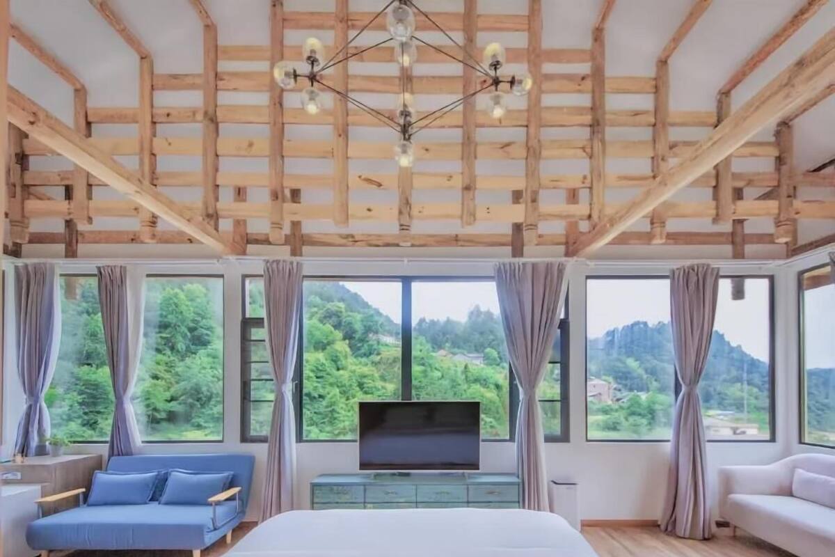 Ein Schlafzimmer mit Bett, Fernseher, Sofa und großen Fenstern mit Blick auf Grünflächen.