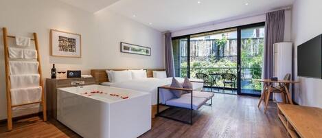 A modern hotel room with a large bed, a separate seating area with a bathtub, a balcony with a view of greenery, and a flat-screen TV.