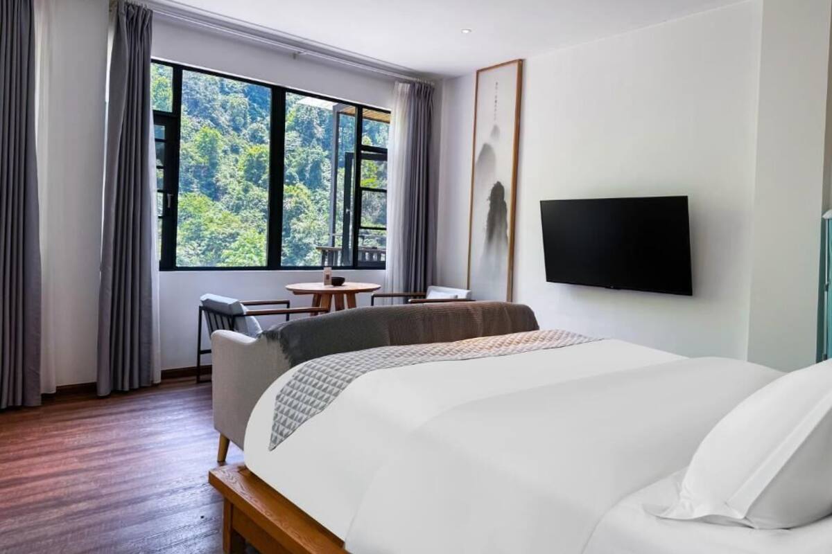 Ein Hotelzimmer mit einem großen Bett, einem Schreibtisch, einem Sessel, einem Fernseher und einem Fenster mit Blick auf Bäume.