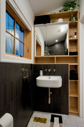 Een badkamer met een witte wasbak, donkere tegels, een raam met houten kozijnen en een spiegel boven de wasbak.