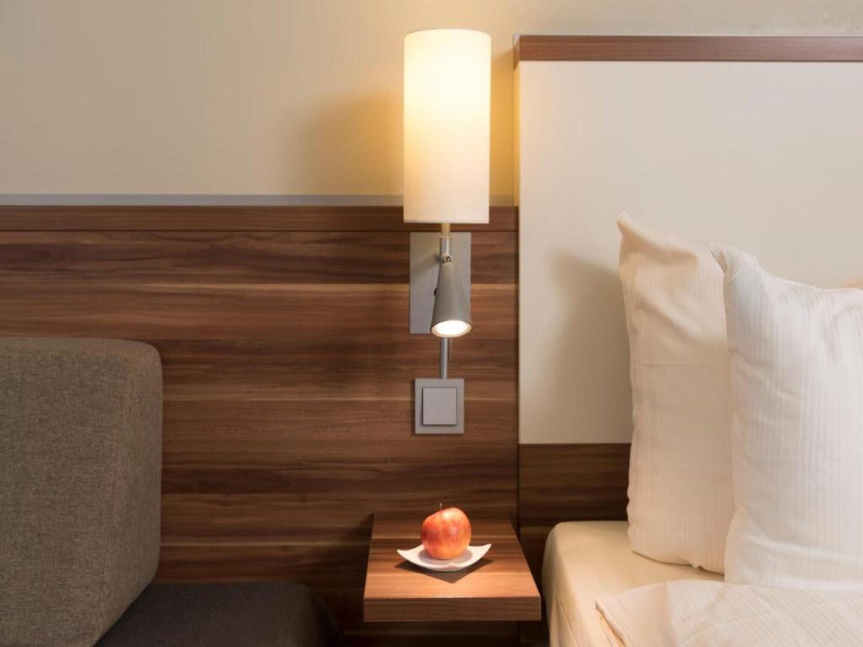 A hotel room with a wooden headboard, a bedside table with a lamp, a pillow, and a fruit on the table.